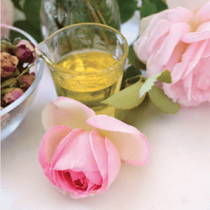A vibrant pink rose, a bowl of dried rose petals, and a glass jug of golden essential oil on a white surface, with a blooming rose in the background.