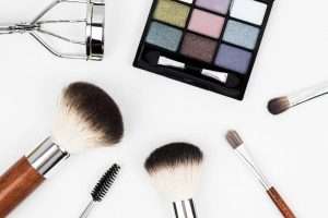 A palette of eyeshadow in a black box and a set of makeup brushes, accompanied by an eyelash curler, are arranged on a white background in an overhead view.