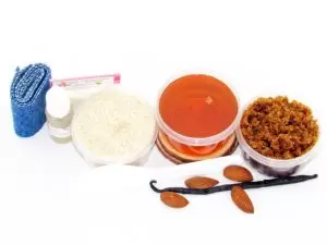 The home sugaring hair removal starter kit comes with a 2 oz tub of soft sugaring wax, a tub of Almond Vanilla Brown Sugar Body Scrub, and a tub of Colloidal Oatmeal Brown Sugar Dry Body Scrub, a small bottle of pure aloe vera, a pouch of cornstarch, denim strips, an applicator, and the pristine white surface is garnished with almonds and a vanilla bean.