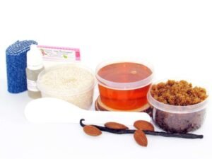 The home sugaring hair removal starter kit comes with a 2 oz tub of soft sugaring wax, a tub of Almond Vanilla Brown Sugar Body Scrub, and a tub of Colloidal Oatmeal Brown Sugar Dry Body Scrub, a small bottle of pure aloe vera, a pouch of cornstarch, denim strips, an applicator, and the pristine white surface is garnished with almonds and a vanilla bean.