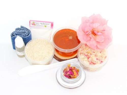The home sugaring hair removal starter kit comes with a 2 oz tub of soft sugaring wax, a tub of Pink Rose Petal Sugar Body Scrub, and a tub of Colloidal Oatmeal Brown Sugar Dry Body Scrub, a small bottle of pure aloe vera, a pouch of cornstarch, denim strips, an applicator, and a glass bowl garnished with a pink rose bloom.