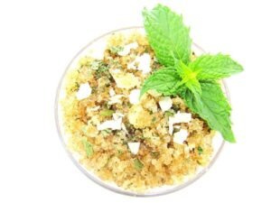 A glass bowl of JBHomemade Sugaring and Skin Care's Natural Peppermint Coconut Sugar Body Scrub with sprinkles of coconut shavings, dried mint and a fresh mint sprig on top.
