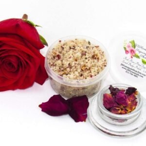 Discover a 2 oz tub of exfoliating cane sugar scrub, enriched with aromatic red rose petals, offers a calming and opulent skin treatment. The scrub's tub is centrally placed, with a glass bowl to the right filled with the sugar scrub and crowned by a vivid, fully bloomed red rose. In the foreground, a smaller glass bowl contains dried rose petals, with additional petals strewn across a white backdrop.