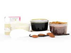 The home sugaring hair removal starter kit comes with a 2 oz tub of firm sugaring paste, a tub of Almond Vanilla Brown Sugar Body Scrub, and a tub of Colloidal Oatmeal Brown Sugar Dry Body Scrub, a small bottle of pure aloe vera, a pouch of cornstarch, an applicator, and the pristine white surface is garnished with almonds and a vanilla bean.