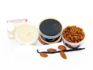 The home sugaring hair removal starter kit comes with a 2 oz tub of firm sugaring paste, a tub of Almond Vanilla Brown Sugar Body Scrub, and a tub of Colloidal Oatmeal Brown Sugar Dry Body Scrub, a small bottle of pure aloe vera, a pouch of cornstarch, an applicator, and the pristine white surface is garnished with almonds and a vanilla bean.