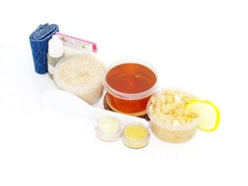 The home sugaring hair removal starter kit comes with a 2 oz tub of soft sugaring wax, a tub of Coconut Lemon Sugar Body Scrub, and a tub of Colloidal Oatmeal Brown Sugar Dry Body Scrub, a small bottle of pure aloe vera, a pouch of cornstarch, an applicator, denim strips, and the pristine white surface is garnished with coconut shavings, lemon zest and slices.