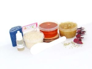 The home sugaring hair removal starter kit comes with a 2 oz tub of soft sugaring wax, a tub of Rosemary Eucalyptus Herbal Sugar Body Scrub, and a tub of Colloidal Oatmeal Brown Sugar Dry Body Scrub, a small bottle of pure aloe vera, a pouch of cornstarch, denim strips, an applicator, and the pristine white surface is garnished with sprinkled dried rosemary and eucalyptus sprigs.