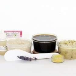 The home sugaring hair removal starter kit comes with a 2 oz tub of firm sugaring paste, a tub of Lavender Lemon Herbal Sugar Body Scrub, and a tub of Colloidal Oatmeal Brown Sugar Dry Body Scrub, a small bottle of pure aloe vera, a pouch of cornstarch, an applicator, and the pristine white surface is garnished with a lavender sprig and lemon zest.