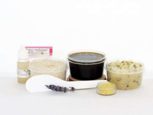 The home sugaring hair removal starter kit comes with a 2 oz tub of firm sugaring paste, a tub of Lavender Lemon Herbal Sugar Body Scrub, and a tub of Colloidal Oatmeal Brown Sugar Dry Body Scrub, a small bottle of pure aloe vera, a pouch of cornstarch, an applicator, and the pristine white surface is garnished with a lavender sprig and lemon zest.
