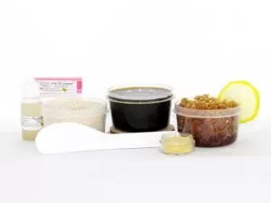 The home sugaring hair removal starter kit comes with a 2 oz tub of firm sugaring paste, a tub of Lemon Zest Sugar Body Scrub, and a tub of Colloidal Oatmeal Brown Sugar Dry Body Scrub, a small bottle of pure aloe vera, a pouch of cornstarch, an applicator, and the pristine white surface is garnished with a lemon zest and slices.