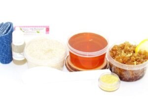 The home sugaring hair removal starter kit comes with a 2 oz tub of soft sugaring wax, a tub of Lemon Zest Sugar Body Scrub, and a tub of Colloidal Oatmeal Brown Sugar Dry Body Scrub, a small bottle of pure aloe vera, a pouch of cornstarch, denim strips, an applicator, and the pristine white surface is garnished with a lemon zest and slices.