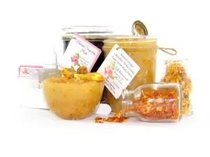 A collection of skincare products including an 8 oz mason jar of sugaring paste and Orange Calendula Herbal Sugar Scrub, accompanied by smaller jars of calendula petals and dried orange zest on a white background.