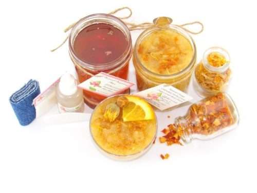 A collection of skincare products including an 8 oz mason jar of sugaring wax and Orange Calendula Herbal Sugar Scrub, the included applicator, bottle of pure aloe vera, denims strips and accompanied by smaller jars of calendula petals and dried orange zest on a white background.
