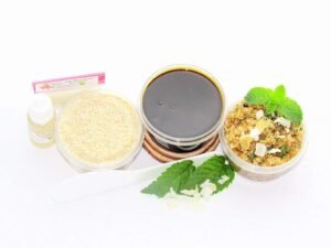 The home sugaring hair removal starter kit comes with a 2 oz tub of firm sugaring paste, a tub of Peppermint Coconut Sugar Body Scrub, and a tub of Colloidal Oatmeal Brown Sugar Dry Body Scrub, a small bottle of pure aloe vera, a pouch of cornstarch, an applicator, and the pristine white surface is garnished with coconut shavings and fresh mint sprigs.