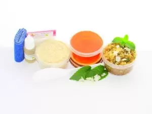 The home sugaring hair removal starter kit comes with a 2 oz tub of soft sugaring wax, a tub of Peppermint Coconut Sugar Body Scrub, and a tub of Colloidal Oatmeal Brown Sugar Dry Body Scrub, a small bottle of pure aloe vera, a pouch of cornstarch, denim strips, an applicator, and the pristine white surface is garnished with coconut shavings and fresh mint sprigs.