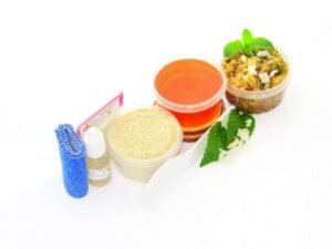 The home sugaring hair removal starter kit comes with a 2 oz tub of soft sugaring wax, a tub of Peppermint Coconut Sugar Body Scrub, and a tub of Colloidal Oatmeal Brown Sugar Dry Body Scrub, a small bottle of pure aloe vera, a pouch of cornstarch, denim strips, an applicator, and the pristine white surface is garnished with coconut shavings and fresh mint sprigs.