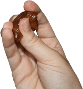 A person holding a ball of sugaring paste for hair removal, demonstrating the ideal size and texture. The photo features a close-up of fingers holding a small ball of sugaring paste. The sugaring paste is dark brown and glossy, indicating its sticky consistency. The size of the sugaring paste ball is ideal, fitting comfortably between two fingers. The background is softly blurred, drawing attention to the sugaring paste itself.