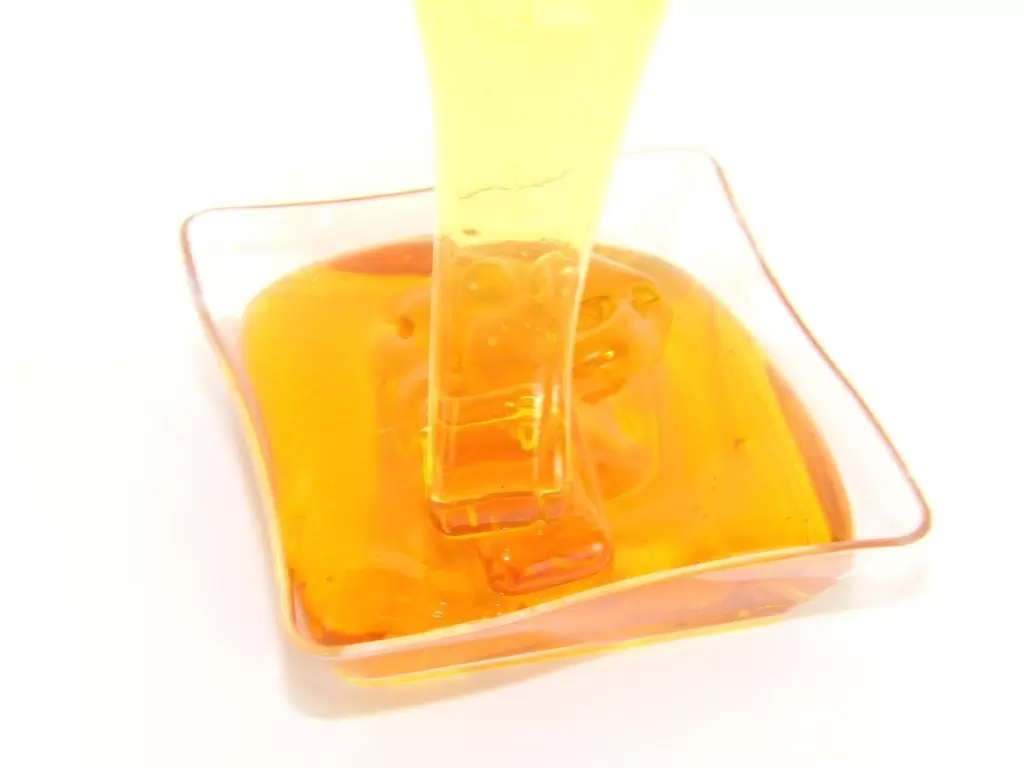 A close-up photograph captures the captivating flow of golden, translucent sugaring wax as it elegantly cascades from a tilted container, accentuating the wax's smooth texture, rich amber hue, and lustrous shine.