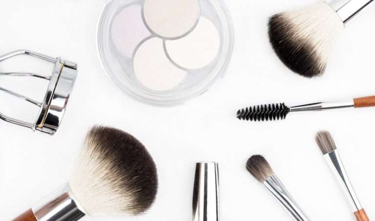 A palette of eyeshadow in a white box and a set of makeup brushes, accompanied by an eyelash curler, are arranged on a white background in an overhead view.