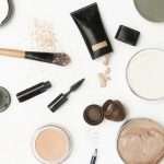 Overhead view of a makeup desk or vanity counter with scattered bottles, tins, powders, mascara, and foundation brushes