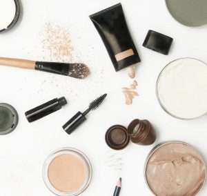 Overhead view of a makeup desk or vanity counter with scattered bottles, tins, powders, mascara, and foundation brushes