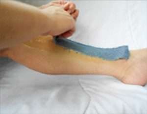 A close-up screenshot from a video which depicts sugaring hair removal using denim strips. The screenshot captures the moment when the denim strip is about to be swiftly removed from the lower inside calf, revealing silky smooth skin.