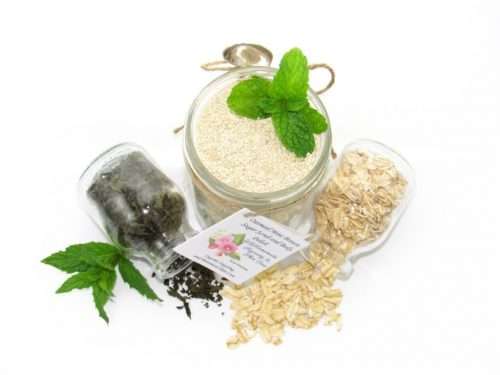 Discover an 8 oz jar of gentle exfoliating dry body scrub, ideal for sensitive skin, featuring colloidal oatmeal, fresh and dried mint, and brown sugar. Accompanying the scrub are glass bottles of oats and dried mint, lying on their sides with their contents artfully spilled on a white table, complemented by sprigs of bright green fresh mint.