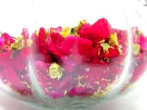 Red rose petals and chamomile flowers being mixed together in a clear glass jar.