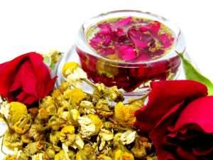 The infused oil in a glass jar surrounded by abundant piles of fresh red roses and dried chamomile.