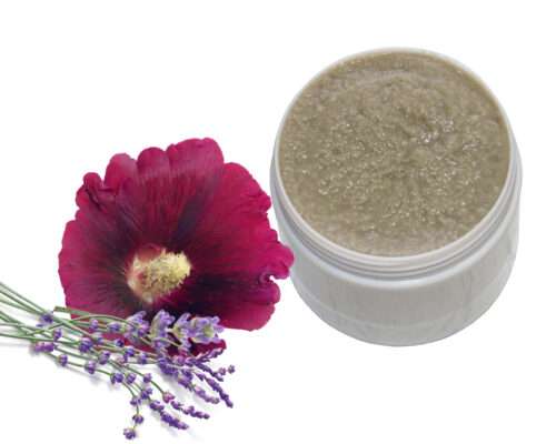 A dark pink hollyhock flower and some lavender sprigs are in front of an open jar of Lavender Hollyhock & Aloe Facial Mask. The jar shows the natural clay mask that hydrates the skin with garden-fresh ingredients. The jar is 2 oz in size and has an angled view.