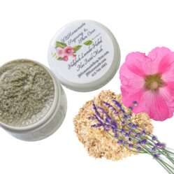 A bird's-eye view of some lavender sprigs on a heap of marshmallow root, beside a pink hollyhock flower. Next to the ingredients, there is a jar of Lavender Hollyhock & Aloe Facial Mask | Natural Clay Facial Mask for Radiant Skin | Handcrafted with Garden-Fresh Ingredients | Hydrating 2 oz. The mask can be seen inside the jar, and the lid is placed next to it, showing the label on the top of the jar.