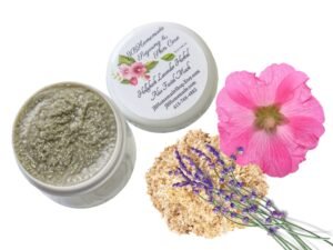 A bird's-eye view of some lavender sprigs on a heap of marshmallow root, beside a pink hollyhock flower. Next to the ingredients, there is a jar of Lavender Hollyhock & Aloe Facial Mask | Natural Clay Facial Mask for Radiant Skin | Handcrafted with Garden-Fresh Ingredients | Hydrating 2 oz. The mask can be seen inside the jar, and the lid is placed next to it, showing the label on the top of the jar.