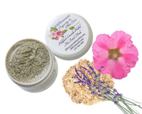 A bird's-eye view of some lavender sprigs on a heap of marshmallow root, beside a pink hollyhock flower. Next to the ingredients, there is a jar of Lavender Hollyhock & Aloe Facial Mask | Natural Clay Facial Mask for Radiant Skin | Handcrafted with Garden-Fresh Ingredients | Hydrating 2 oz. The mask can be seen inside the jar, and the lid is placed next to it, showing the label on the top of the jar.