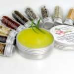 Small glass jars containing ground cloves, turmeric, ginger, St. John’s Wort, frankincense, sandalwood, patchouli, lavender, ylang-ylang extra, and rosemary, arranged around the Comfort Harmony Salve tin.