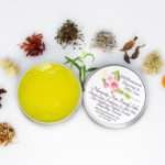 A 2 oz tin of Comfort Harmony Salve opened to reveal the golden, oil-infused herbal salve, surrounded by its natural ingredients like cloves, turmeric, and lavender on a white background.