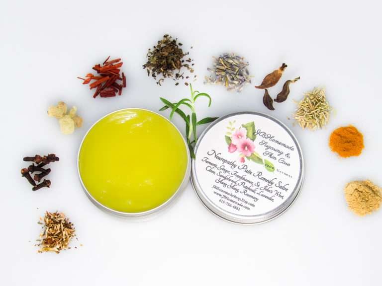 A 2 oz tin of Comfort Harmony Salve opened to reveal the golden, oil-infused herbal salve, surrounded by its natural ingredients like cloves, turmeric, and lavender on a white background.