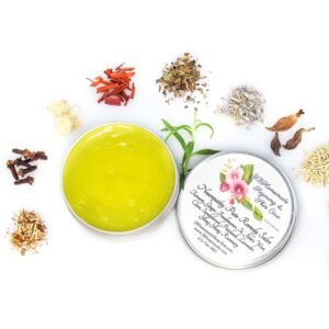 A 2 oz tin of Comfort Harmony Salve opened to reveal the golden, oil-infused herbal salve, surrounded by its natural ingredients like cloves, turmeric, and lavender on a white background.