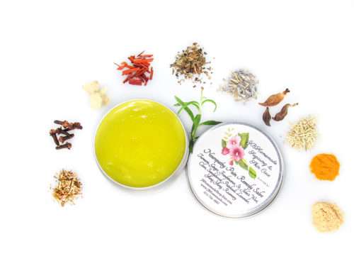 A 2 oz tin of Comfort Harmony Salve opened to reveal the golden, oil-infused herbal salve, surrounded by its natural ingredients like cloves, turmeric, and lavender on a white background.