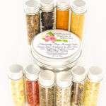 Small glass jars containing ground cloves, turmeric, ginger, St. John’s Wort, frankincense, sandalwood, patchouli, lavender, ylang-ylang extra, and rosemary, arranged around the Comfort Harmony Salve tin.
