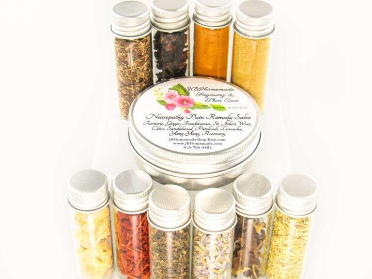 Small glass jars containing ground cloves, turmeric, ginger, St. John’s Wort, frankincense, sandalwood, patchouli, lavender, ylang-ylang extra, and rosemary, arranged around the Comfort Harmony Salve tin.