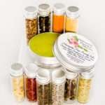 Small glass jars containing ground cloves, turmeric, ginger, St. John’s Wort, frankincense, sandalwood, patchouli, lavender, ylang-ylang extra, and rosemary, arranged around the Comfort Harmony Salve tin.