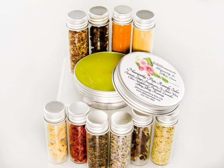 Small glass jars containing ground cloves, turmeric, ginger, St. John’s Wort, frankincense, sandalwood, patchouli, lavender, ylang-ylang extra, and rosemary, arranged around the Comfort Harmony Salve tin.