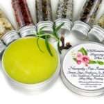 Small glass jars containing ground cloves, turmeric, ginger, St. John’s Wort, frankincense, sandalwood, patchouli, lavender, ylang-ylang extra, and rosemary, arranged around the Comfort Harmony Salve tin.