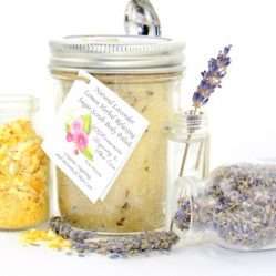 Discover an 8 oz jar of calming cane sugar scrub, a blend of relaxing lavender and invigorating lemon, ideal for a soothing experience. The sugar scrub jar is placed centrally, adorned with dried lemon zest and a lemon slice on top. Flanking the scrub jar are two smaller jars: one brimming with dried lavender buds, the other with lemon zest, both casually tipped over, their contents artfully scattered on a pristine white surface. A delicate lavender sprig rests in the tiniest jar, positioned to the right of the sugar scrub.