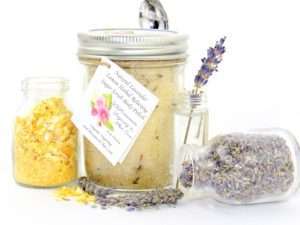 Discover an 8 oz jar of calming cane sugar scrub, a blend of relaxing lavender and invigorating lemon, ideal for a soothing experience. The sugar scrub jar is placed centrally, adorned with dried lemon zest and a lemon slice on top. Flanking the scrub jar are two smaller jars: one brimming with dried lavender buds, the other with lemon zest, both casually tipped over, their contents artfully scattered on a pristine white surface. A delicate lavender sprig rests in the tiniest jar, positioned to the right of the sugar scrub.
