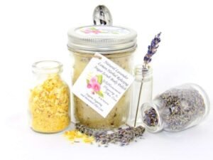 Discover an 8 oz jar of calming cane sugar scrub, a blend of relaxing lavender and invigorating lemon, ideal for a soothing experience. The sugar scrub jar is placed centrally, adorned with dried lemon zest and a lemon slice on top. Flanking the scrub jar are two smaller jars: one brimming with dried lavender buds, the other with lemon zest, both casually tipped over, their contents artfully scattered on a pristine white surface. A delicate lavender sprig rests in the tiniest jar, positioned to the right of the sugar scrub.