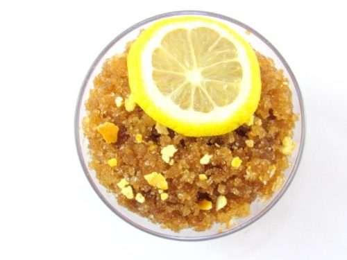 A glass bowl of JBHomemade Sugaring and Skin Care's Natural Lemon Zest Citrus Sugar Body Scrub adorned with sprinkles of dried lemon zest and a slice of lemon.