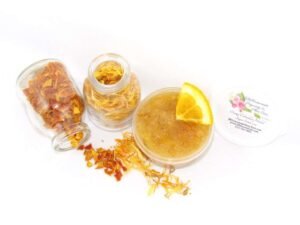 A 2 oz tub of vibrant citrus cane sugar scrub, infused with orange and calendula, is designed to enhance a radiant glow. The sugar scrub tub is placed centrally, flanked by two smaller jars—one filled with dried orange zest, the other with dried calendula—both spilling their contents onto a white backdrop. The tub brims with the sugar scrub, adorned with calendula petals and crowned with a fresh orange slice.