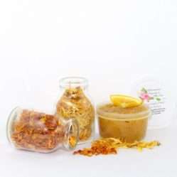 A 2 oz tub of vibrant citrus cane sugar scrub, infused with orange and calendula, is designed to enhance a radiant glow. The sugar scrub tub is placed centrally, flanked by two smaller jars—one filled with dried orange zest, the other with dried calendula—both spilling their contents onto a white backdrop. The tub brims with the sugar scrub, adorned with calendula petals and crowned with a fresh orange slice.