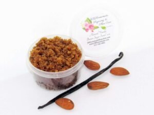 Centered is a 2 oz tub of natural brown sugar body scrub, enriched with almond, honey, and vanilla. In the foreground lies scattered almonds and a whole vanilla bean.